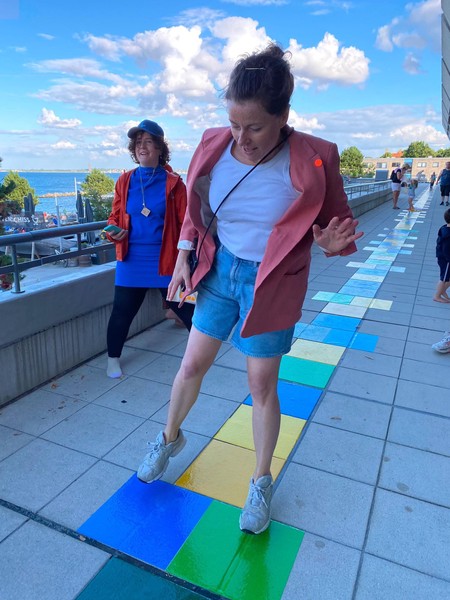 Das Bild zeigt zwei Frauen. Die eine im Vordergrund spingt gerade entlang eines sehr langen Hüpfspiels. Dies kann man erkennen, da die punkten Kästen bis weit in den Bildhintergrund reichen. Im Hintergrund sind weitere Personen auf der Hüpfstrecke zu erkennen. Die zweite Frau steht kurz dahinter neben der Strecke mit sportlicher Kleidung, Basecap und einer Art Stoppuhr in der Hand. Beide Frauen lächeln. Der Himmel ist blau mit wenigen Wolken. Im Hintergrund kann man das Meer sehen.