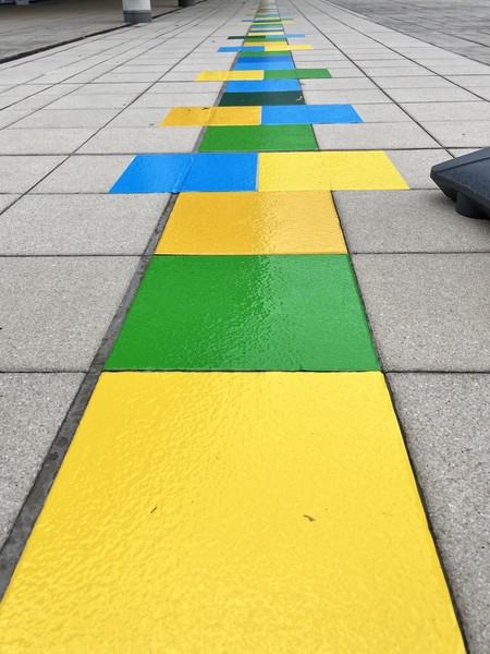 Das Bild zeigt ein in gelb, orange, blau und grün auf den Boden aufgebrachtes überlanges Hüpfspiel in Nahaufnahme.