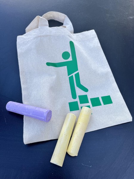 Das Bild zeigt einen Jutebeutel mit dem oben beschriebenen Hüpfspielpiktogramm in grün. Auf dem Beutel liegen drei Straßenmalkreidestifte.