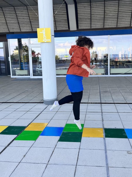 Das Bild zeigt eine erwachsene Person in der Luft über dem oben beschriebenen überlangen Hüpfspiel. Im Hintergrund ist ein gelbes Schild, das auch das oben beschriebene olympische Piktogramm enthält.