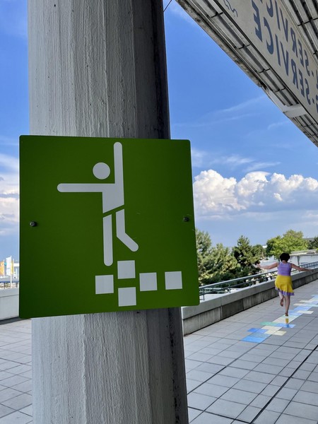 Das Bild zeigt ein grünes Schild im Vordergrund, das eine Art Sportpiktogramm enthält. Dieses an die olympischen Piktogramme von 1972 erinnernde Symbol zeigt eine Figur, die ein Hüpfspiel ausübt. Im Hintergrund ist eine Person zu sehen, die ein buntes auf dem Boden angebrachtes Hüpfspiel ausübt.