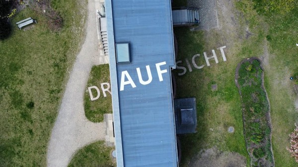 Das Bild zeigt eine Drohnenansicht eines Gebäudedaches mit Rasenflächen und Wegen auf beiden Seiten. Schräg rüber steht das Wort „DRAUFSICHT“