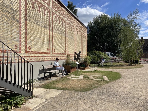 Das Bild zeigt ein Backsteingebäude im linken Bildrand. Davor auf einer Bank sitzt eine weiß gekleidete Person. Zu ihren Füßen auf dem Boden kann man über Kopf die Buchstaben D und R lesen.