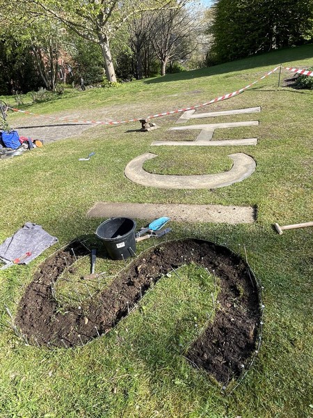 Das Bild zeigt eine Rasenfläche, die von einem Baustellenabsperrband eingegrenzt ist. Auf dem Boden steht das Wort SICHT. Man kann sehen, dass das S in den Rasen gegraben ist. Die Buchstaben I, C, H und T sind bereits mit Beton ausgegossen.