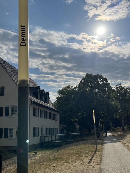 Das Bild zeigt eine schmale Leuchte bei Tag, die nicht eingeschaltet ist, dahinter Bäume und ein Gebäude. Auf ihr steht in schwarzen Buchstaben: Demut