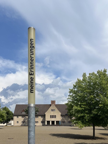 Das Bild zeigt eine schmale Leuchte bei Tag, die nicht eingeschaltet ist, dahinter ein Baum, ein großer Platz und ein großes Gebäude. Auf ihr steht in schwarzen Buchstaben: meine Erinnerungen