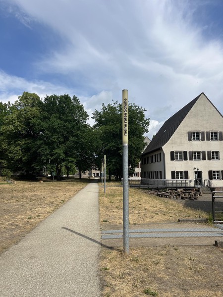 Das Bild zeigt eine schmale Leuchte bei Tag, die nicht eingeschaltet ist, dahinter Bäume und ein Gebäude. Auf ihr steht in schwarzen Buchstaben: meine roten Schuhe