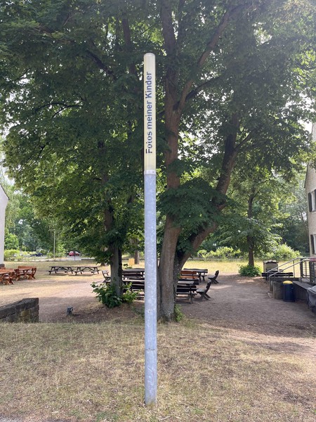 Das Bild zeigt eine schmale Leuchte bei Tag, die nicht eingeschaltet ist, dahinter Bäume und ein Gebäude. Auf ihr steht in schwarzen Buchstaben: Fotos meiner Kinder