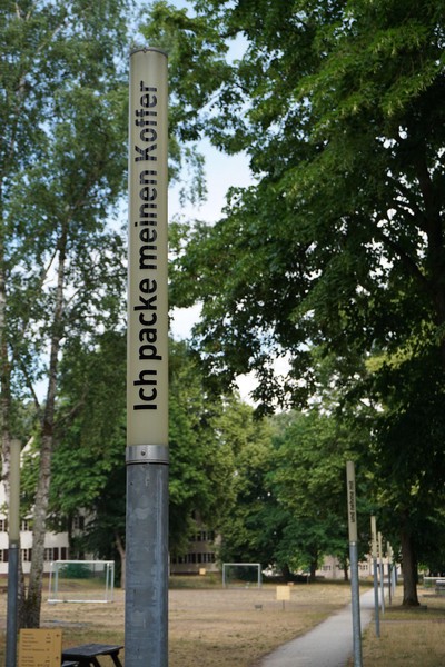 Das Bild zeigt ein Gelände mit Bäumen und Gebäuden, sowie Lampen, die an einem Weg stehen. Die Lampen sind nicht eingeschaltet, weil es Tag ist. Sie haben eine schmale schlichte Form, bestehen unten aus einem Metallrohr und oben aus einem cremeweißen Kunststoffzylinder. Auf der Lampe im Vordergrund steht in schwarzen Buchstaben: „Ich packe meinen Koffer“.