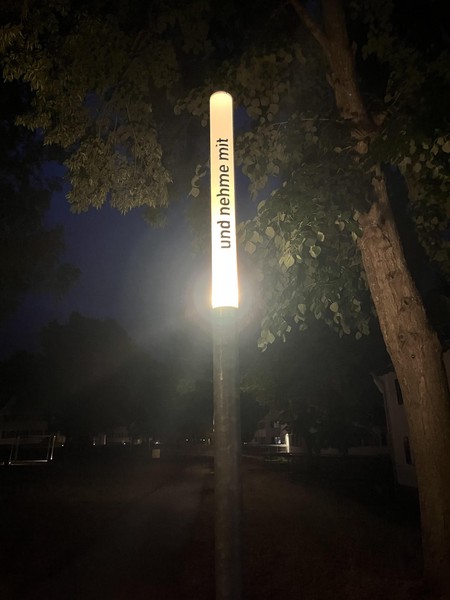 Das Bild zeigt eine schmale Leuchte bei Nacht, die eingeschaltet ist. Auf ihr steht in schwarzen Buchstaben: … und nehme mit 