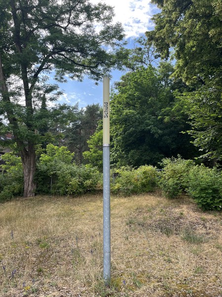 Das Bild zeigt eine schmale Leuchte bei Tag, die nicht eingeschaltet ist, dahinter Bäume. Auf ihr steht in schwarzen Buchstaben: Schuld