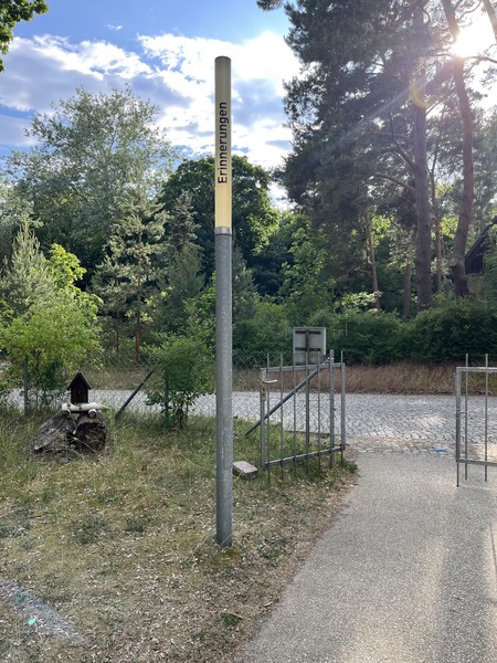 Das Bild zeigt eine schmale Leuchte bei Tag, die nicht eingeschaltet ist, dahinter Bäume und ein Eingangstor. Auf ihr steht in schwarzen Buchstaben: Erinnerungen