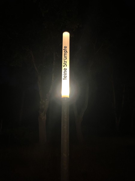 Das Bild zeigt eine schmale Leuchte bei Nacht, die eingeschaltet ist. Auf ihr steht in schwarzen Buchstaben: feine Strümpfe