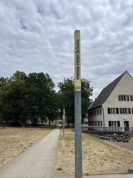 Das Bild zeigt eine schmale Leuchte bei Tag, die nicht eingeschaltet ist, dahinter Bäume und ein Gebäude. Auf ihr steht in schwarzen Buchstaben: meine roten Schuhe