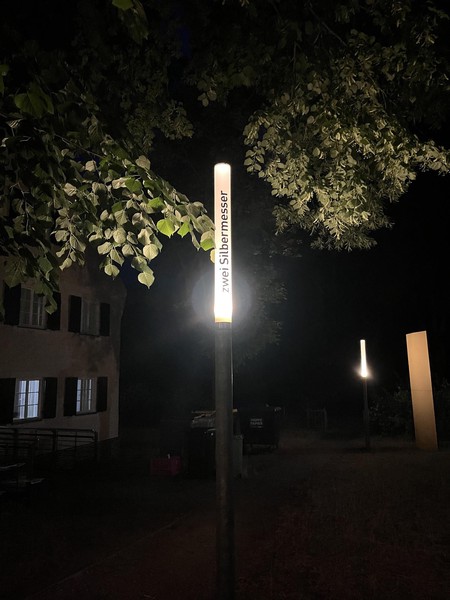 Das Bild zeigt eine schmale Leuchte bei Nacht, die eingeschaltet ist. Auf ihr steht in schwarzen Buchstaben: zwei Silbermesser 