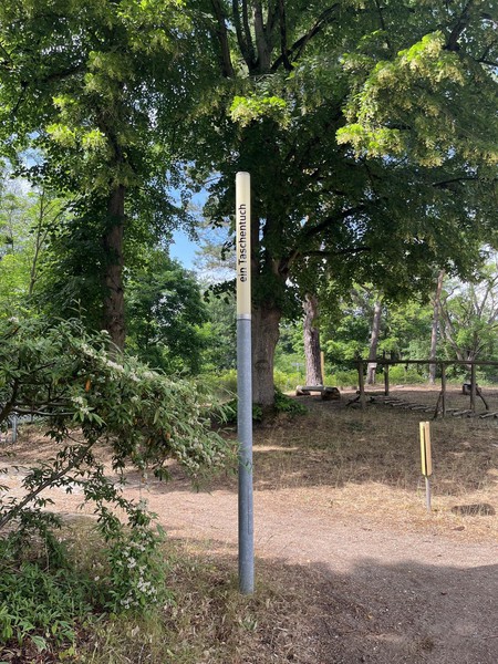 Das Bild zeigt eine schmale Leuchte bei Tag, die nicht eingeschaltet ist, dahinter Bäume. Auf ihr steht in schwarzen Buchstaben: ein Taschentuch