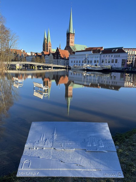 Das Bild zeigt im Hintergrund eine Kirche hinter einem Gewässer. Im vorderen Bereich erkennt man eine Bronzetafel, die ein Relief andeutet.