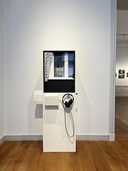 Das Bild zeigt ein schwarz-weiß gestaltetes Objekt, das einem Fenster nachempfunden ist. Man erkennt die Beschläge. Davor steht ein Chanukka-Leuchter in weiß und links hängt ein schwarz-weiß gemusterter Vorhang. Das Objekt befindet sich auf einem weißen Sockel, an dem sich weitere weiße Kästen und ein Kopfhörer befinden. Im rechten Bildrand kann man einen weiteren Ausstellungsraum erkennen.