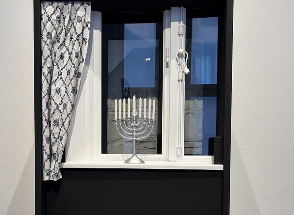 Das Bild zeigt ein schwarz-weiß gestaltetes Objekt, das einem Fenster nachempfunden ist. Man erkennt die Beschläge. Davor steht ein Chanukka-Leuchter in weiß und links hängt ein schwarz-weiß gemusterter Vorhang.