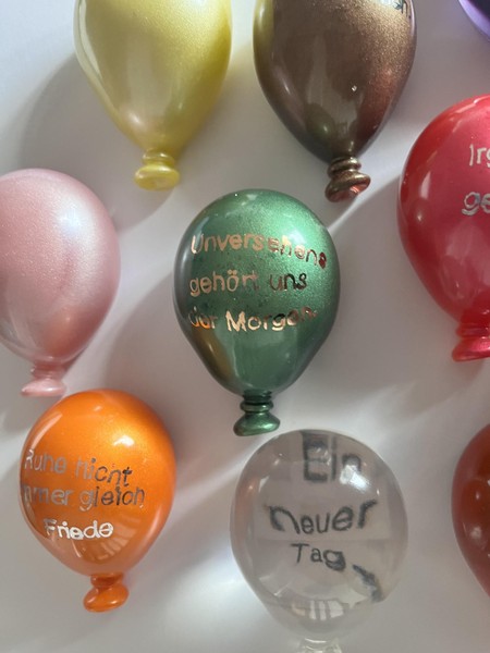 Das Bild zeigt eine Detailaufnahme von acht der kleinen Luftballons. Auf mehreren kann man Textfragmente erkennen. Lesbar sind die Texte: Unversehens gehört uns der Morgen, Ein neuer Tag, Ruhe nicht immer gleich Friede