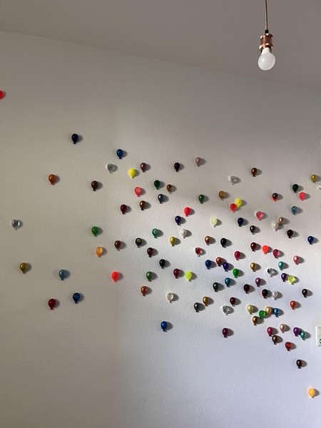 Das Bild zeigt eine Zimmerwand, erkennbar an Glühbirne am oberen Bildrand. Die ganze Wand ist voller bunter kleiner Luftballons.