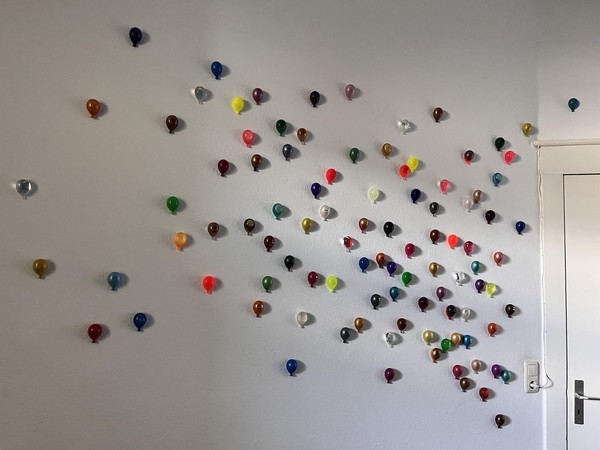 Das Bild zeigt eine Zimmerwand, erkennbar an einer Tür am rechten Bildrand. Die ganze Wand ist voller bunter kleiner Luftballons.