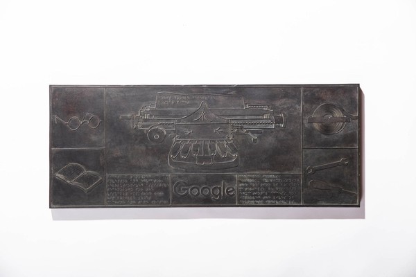 Das Bild zeigt eine Bronzetafel mit grafischen Darstellungen. Es ist in acht unterschiedlich große Felder aufgeteilt. Zentral in der Mitte am größten im Oberen Bereich ist eine Art Schreibmaschine zu sehen, die als Brailleschreibmaschine an den sechs Tasten und dem eingespannten Papier zu erkennen ist, auf dem ein Satz in Brailleschrift steht. Auf der linken Seite ist oben eine Brille und unten ein Buch zu erkennen, auf der rechten Seite oben eine Art Spule und unten jeweils ein Schraubenschlüssel und ein Schraubendreher. Der untere mittlere Teil ist noch einmal in drei Bereiche aufgeteilt. Links und recht ist jeweils Brailleschrift zu erkennen, in der Mitte befindet sich das klassische Google Logo.