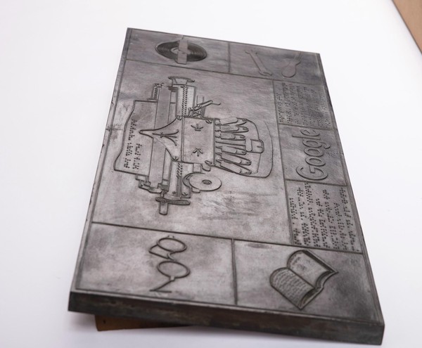 Das Bild zeigt die oben beschriebenen Tafel aus einer anderen Perspektive. Vorne im Bildraum befinden sich Brille und Buch. Durch den Winkel kann man mehr Kontraste erkennen und sehen, dass alle grafischen Elemente taktil erhaben sind.