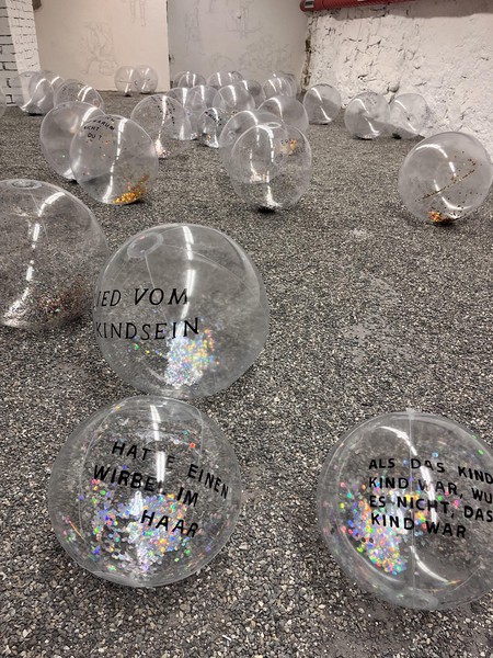 Das Bild zeigt einen Raum mit Kiesboden und weißen Wänden. Das Foto ist ziemlich knapp über dem Boden aufgenommen. Auf dem Boden liegen über 20 transparente Wasserbälle, in deren Innerem Glitter erkennbar ist, der sich gut mit den kleinen Kieselsteinen des Bodens verbindet. Man kann auf den vorderen der drei Bälle schwarze Textfragmente erkennen. Auf einem steht: „Lied vom Kindsein“, auf dem nächsten: „hatte einen Wirbel im Haar“ und auf dem dritten: „Als das Kind, Kind war, wusste es nicht, dass es Kind war“