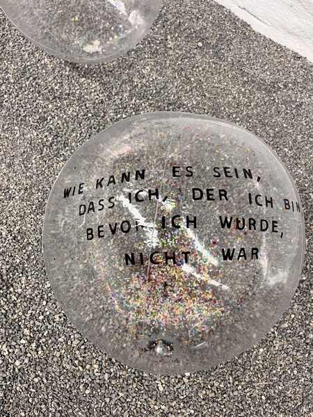 Das Bild ist von oben aufgenommen und ist auf den zuvor beschriebenen Kieselsteinbodengerichtet. Es zeigt einen der beschriebenen transparenten Wasserbälle mit Glitter innendrin. Auf dem Ball steht mit schwarzer Schrift: „Wie kann es sein, dass ich, der ich bin, bevor ich wurde, nicht war“. Ein zweiter Ball ist im Anschnitt zu sehen.