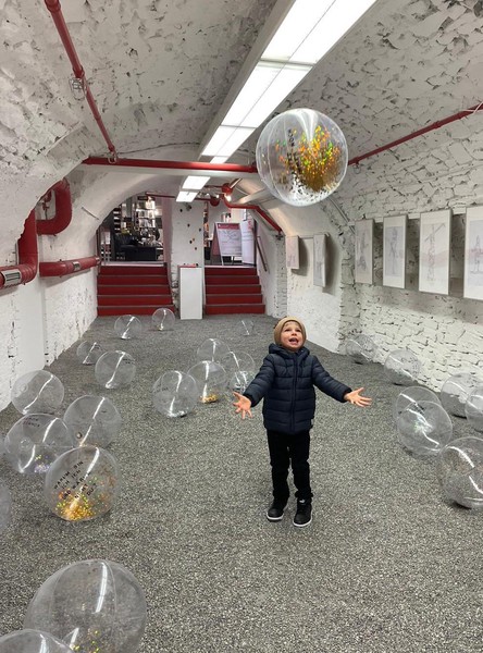 Das Bild zeigt denselben Raum mit Kieselboden und Wasserbällen aus einer anderen Perspektive. Man kann rote Heizungsrohre an den Wänden, auf der rechten Seite gerahmte Bilder und im Hintergund einen Treppenaufgang mit ebenfalls roten Stufen erkennen. Außerdem offenbart die weißgestrichene Backsteindecke, dass es sich um ein Gewölbe handelt. Inmitten der Bälle steht ein kleiner Junge mit Winterkleidung und wirft lachend einen der Bälle in die Höhe.