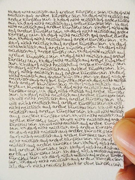 Das Bild zeigt einen Daumen und Zeigefinger in Nahaufnahme, die einen sehr kleinen Zettel halten, wie man an den Größenverhältnissen von Fingern und Papiergröße erkennen kann. Trotz des kleinen Formats ist der Zettel sehr voll geschrieben. Es ist zu erkennen, dass die Handschrift sehr klein gehalten ist. Es steht in über dreißig Zeilen der Satz 'Ich darf nicht neidisch auf andere Künstler sein.' Zwischendrinn wechselt er für ein paar Sätze in 'Ich will nicht neidisch auf andere Künstler sein.' um dann wieder bis zum Schluss anhaltend in 'Ich darf nicht neidisch auf andere Künstler sein.' wechselt.